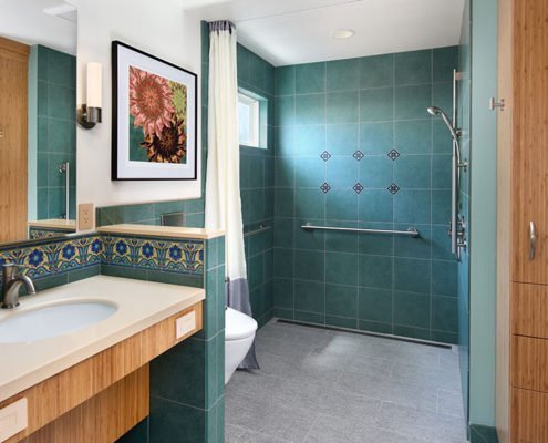 A universal design bathroom inspired by Catalina tiles features a curbless shower with hand railings, a wall-mounted toilet, and a roll-under vanity.