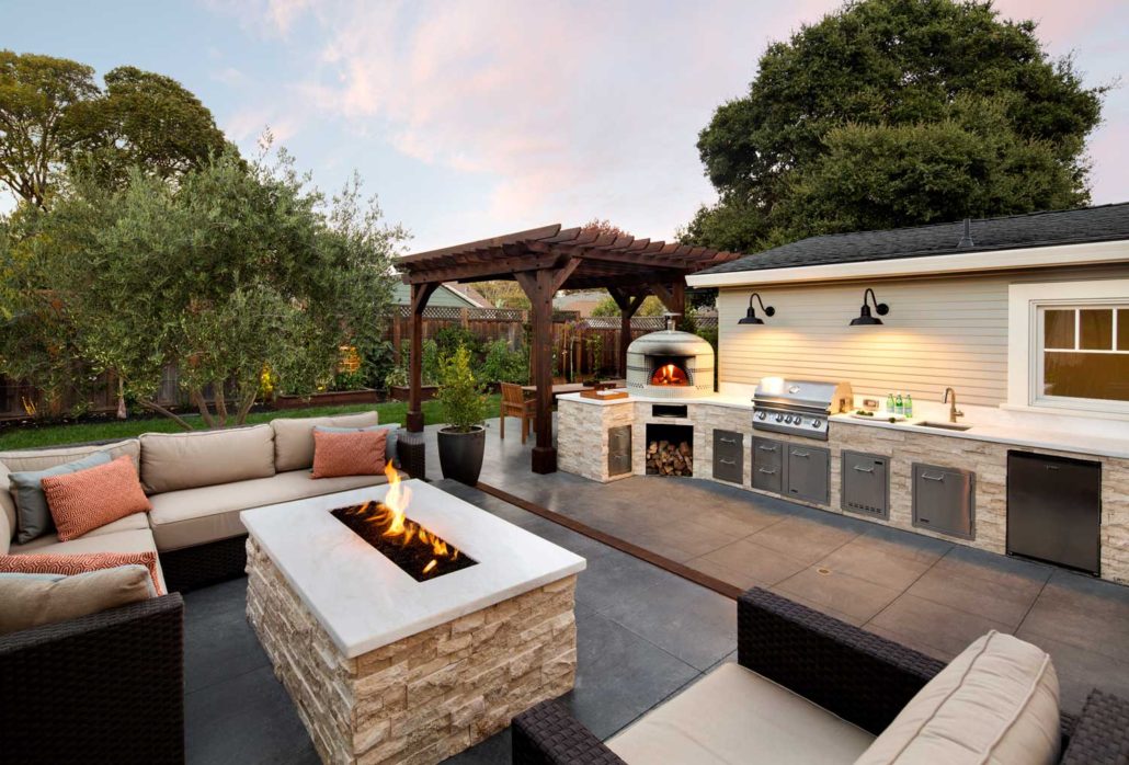 Firepit seating area next to outdoor kitchen with grill and pizza oven
