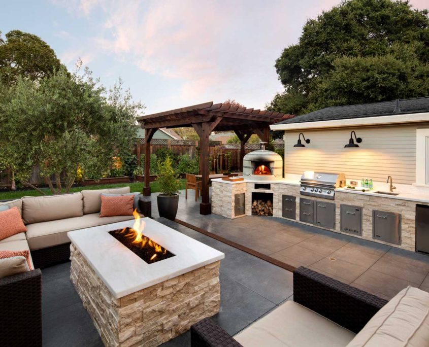 Firepit seating area next to outdoor kitchen with grill and pizza oven