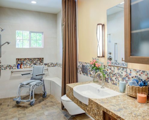 A universal design bathroom with a tiled accent has a roll-under stone vanity, wall-mounted toilet, and curbless shower with a shower wheelchair in it.