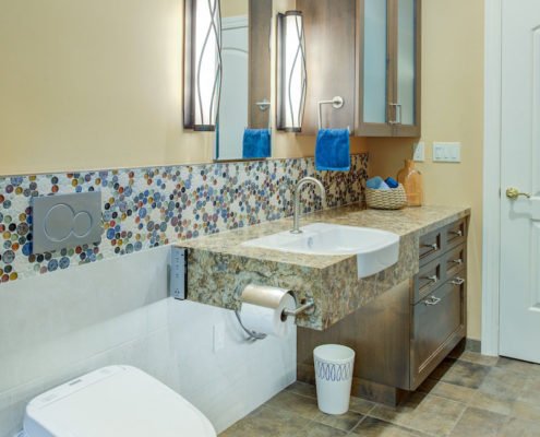 A universal design bathroom with a tiled accent has a roll-under stone vanity and a wall-mounted toilet with a flush plate.