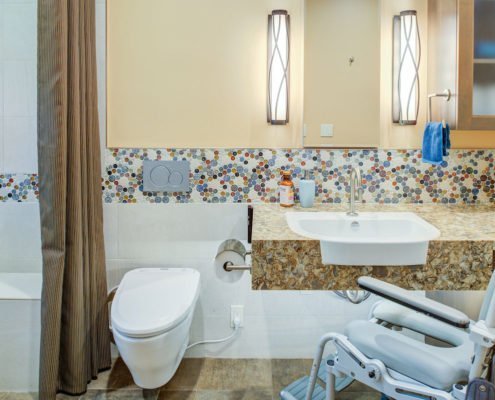 A universal design bathroom with a tiled accent has a roll-under stone vanity, wall-mounted toilet, and curbless shower with a bench.