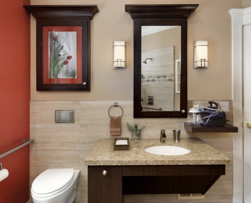 A universal design bathroom has a wall-mounted toilet with a flush plate and roll-under vanity.