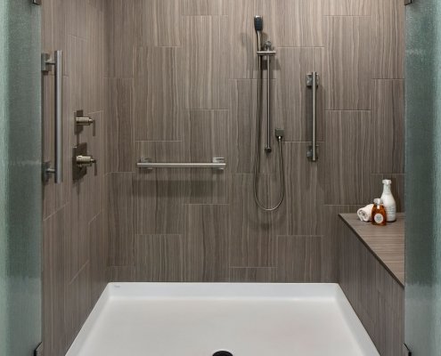A universal design bathroom has a roll-in shower with brushed silver railings, dual shower heads, and a bench.