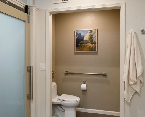 A contemporary universal design bathroom has an extra wide doorway leading to the toilet so it can be accessed by someone in a wheelchair. Brushed metal railings are on the wall to the left of and behind the toilet.