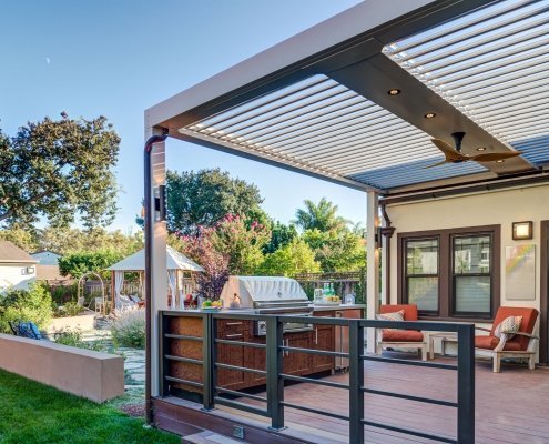 A metal pergola with movable louvers covers an outdoor kitchen and grill.